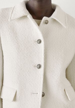 Manteau en laine couleur crème avec une finition texturée, grand col et boutons en argent. Il dispose de poches latérales et d'une coupe ajustée.