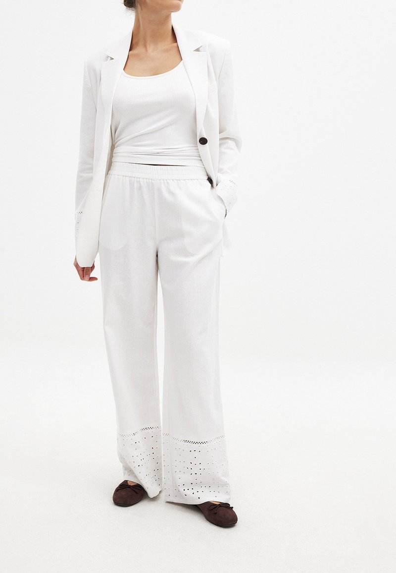 Baltas pritaikytas švarkas užmestas ant baltos prigludusios marškinėlių, derinys su baltais plačiais kelnėmis, kurių apačioje yra iškarpytas raštas. Rudos plokščios bateliai.