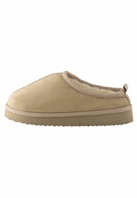 Zueco de ante beige con forro de felpa, de forma redondeada y con bordes en contraste. Presenta una suela de goma texturizada baja y un lazo en la parte trasera.