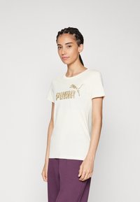 Camiseta de algodón color crema con un logotipo gráfico dorado de "PUMA" en el frente, mangas cortas y un escote redondo.