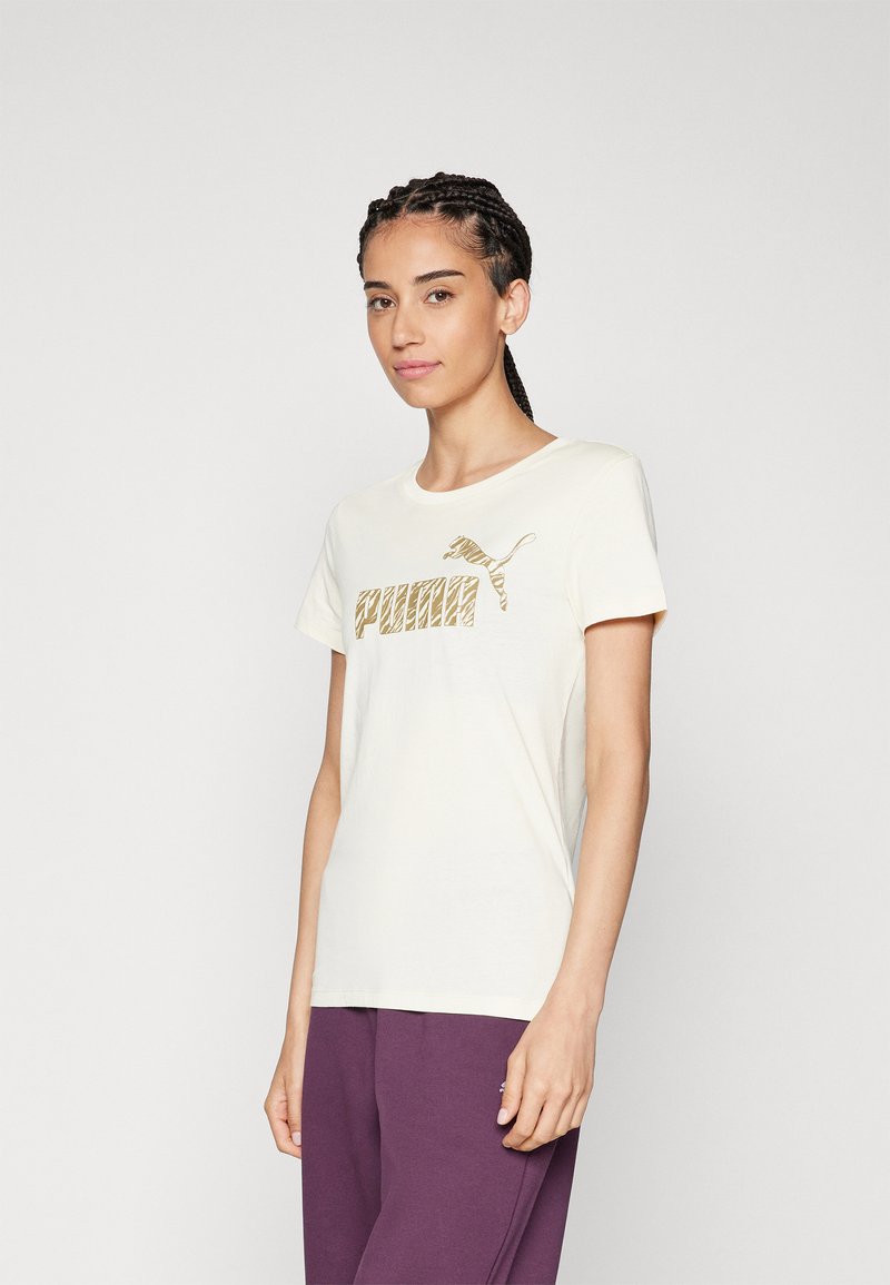 Camiseta de algodón color crema con un logotipo gráfico dorado de "PUMA" en el frente, mangas cortas y un escote redondo.