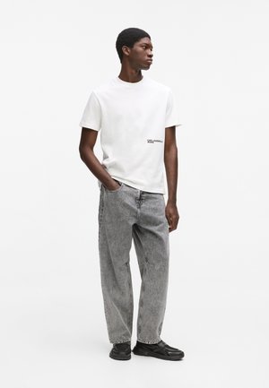 Fiatal férfi fehér Karl Lagerfeld Jeans pólóban, szürke bő szárú farmerral és fekete tornacipővel, egyik kezét zsebében tartva áll.