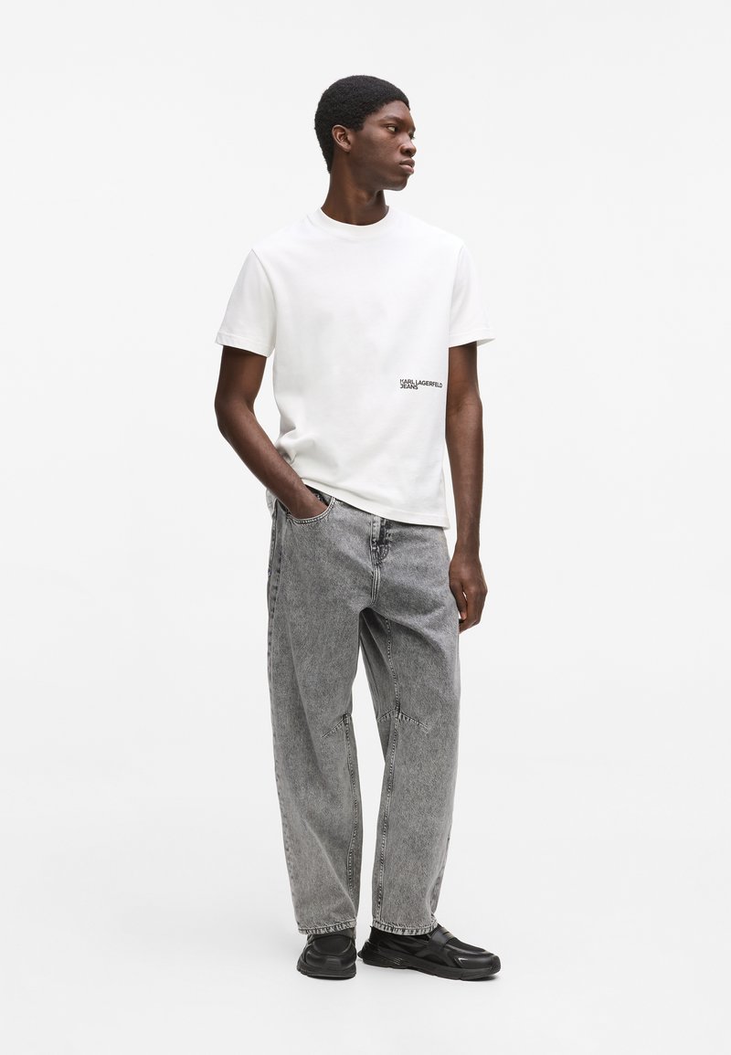 Jeune homme portant un t-shirt blanc Karl Lagerfeld Jeans, un jean gris ample et des baskets noires, debout avec une main dans la poche.