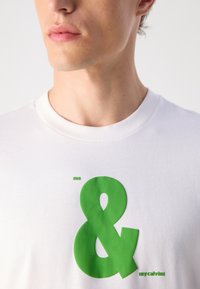 Baltos medvilnės marškinėliai su dideliu žaliu guma užklijuotu ženklu "&" simboliu, o šalia maškinėliuose užrašyta mažesniais žalios spalvos rašmenimis "me" ir "mycalvins".