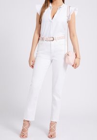 Femme portant un haut sans manches blanc avec un col brodé, un jean taille haute blanc, une ceinture rose, un sac bandoulière rose et des talons roses à lanières.