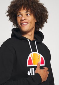 Sudadera negra con un logo colorido que presenta acentos en rojo, naranja y amarillo. Cuerdas de ajuste blancas con puntas metálicas plateadas; textura de tela suave.