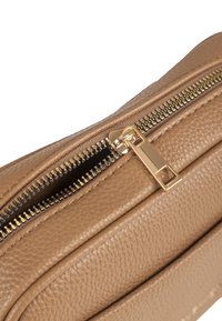 Nahaufnahme einer teilweise geöffneten, hellbraunen, strukturierten Ledertasche mit goldenem Metallreißverschluss und Reißverschlussanhänger.