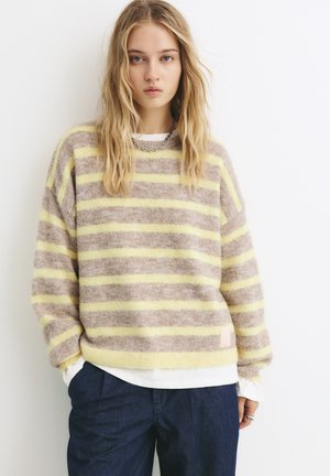 Stribet sweater i beige og gul, lavet af blødt materiale, med en løs pasform og ribbede manchetter, lagdelt over en hvid top. Denim bukser nedenunder.