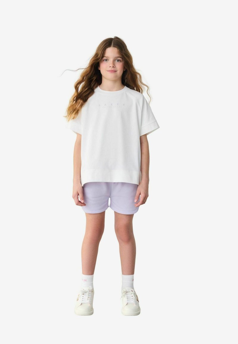 Camiseta básica blanca de gran tamaño con "BAKER" impreso en gris claro, combinada con pantalones cortos lila. El modelo lleva zapatillas blancas y calcetines claros.