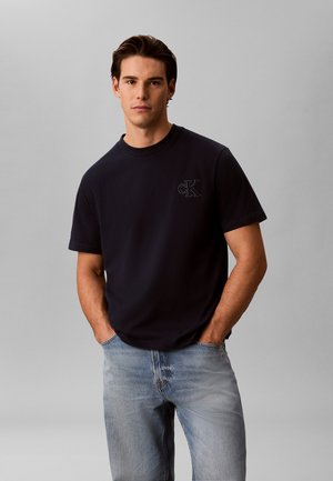 Junger Mann mit dunklen Haaren trägt ein schwarzes CK T-Shirt und hellblaue Jeans, steht mit den Händen in den Taschen vor grauem Hintergrund.