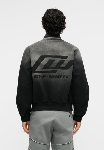 Άνδρας με κατσαρά μαλλιά φοράει μια ξεθωριασμένη μαύρη μπουφάν bomber με μεγάλο λογότυπο "Off-White" στην πλάτη, συνδυασμένη με φαρδιά γκρι παντελόνια.