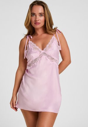 Vrouw die een mouwloze roze satijnen slipjurk draagt met kanten details en strikbandjes, staand tegen een effen grijze achtergrond.