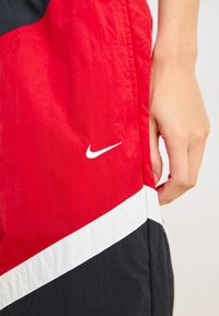 Piros és fekete szélálló dzseki fehér kiegészítőkkel. Jellemzője egy kiemelkedő Nike logó, átlós színblokk és könnyű, texturált felület.