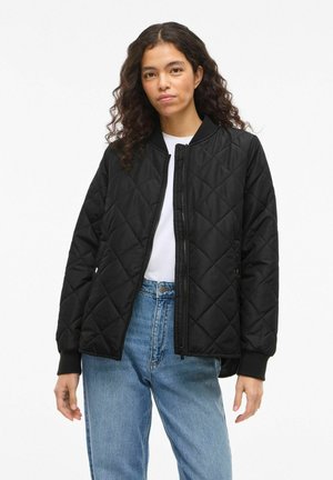 Femme aux cheveux bouclés portant une veste matelassée noire par-dessus une chemise blanche et un jean bleu, debout devant un fond blanc uni.