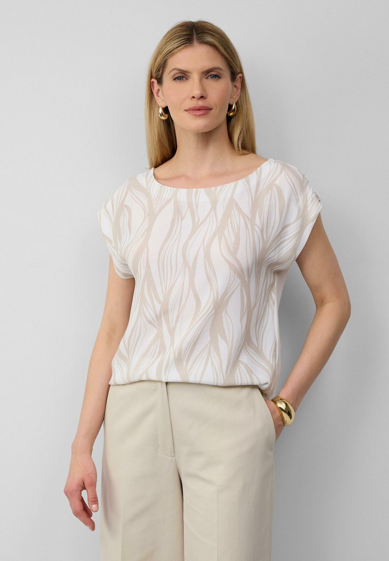 Lichte witte blouse met een vloeiende ontwerp featuring beige bladeren-achtige patronen, korte mouwen en een ronde halslijn, gecombineerd met beige broeken.