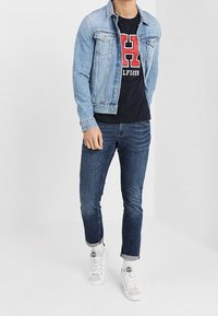 Jeansjacka med ljus tvätt, slim fit mörka jeans, marinblå t-shirt med en röd logotyp, och vita high-top sneakers.