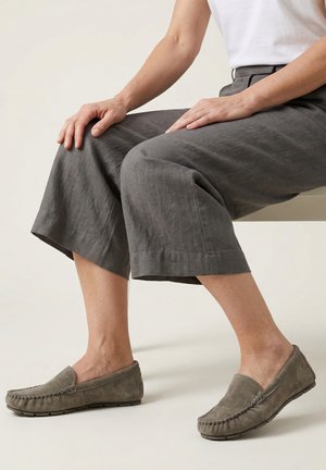 Osoba sedící v šedých krátkých kalhotách a odpovídajících šedých semišových mokasínách, ruce spočívají na stehnech na jednobarevném pozadí.