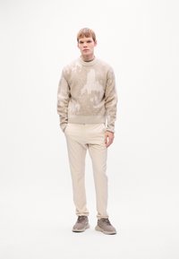Jonge man die staat met zijn handen in zijn zakken, gekleed in een beige gebreid sweater, lichte broek en taupe sneakers tegen een witte achtergrond.