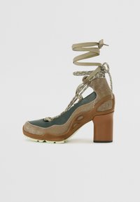 High-heeled shoe with a brown chunky heel, green toe and sides, glittery gold accents, and spiraled laces with a textured finish.