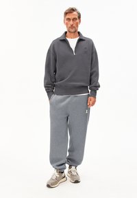 Donkergrijze zip-up sweatshirt met hoge kraag, gecombineerd met lichtgrijze sweatpants met een zijzak. De schoenen zijn grijs en beige.