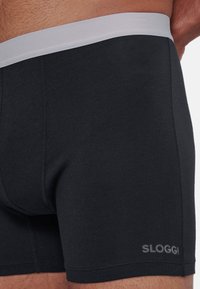 Boxer da uomo in cotone nero con elastico grigio in vita, caratterizzati da cuciture piatte e logo SLOGGI stampato in grigio in basso a sinistra.