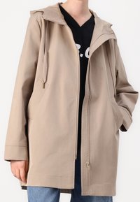 Giacca beige con cappuccio e cerniera frontale, dotata di tasche laterali, vestibilità morbida e dettagli di cuciture sottili. Indossata sopra a una maglia nera.