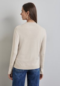 Beige Strickpullover mit Rippenkragen, langen Ärmeln und einer leicht lockeren Passform, kombiniert mit dunkelblauen Jeans.