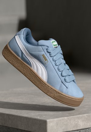 Jasnoniebieski zamszowy sneaker Puma z białym paskiem i gumową podeszwą, unoszący się nad betonową powierzchnią na tle ciemnego tła.