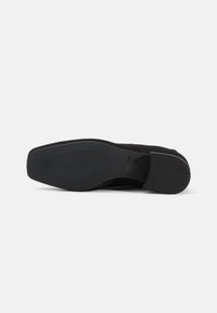 Black suede shoe with a flat design, textured rubber sole, and smooth curved edges, featuring minimal branding on the insole.