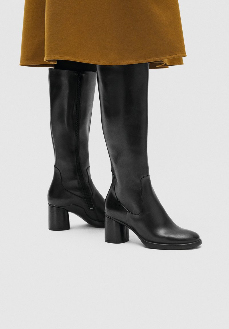 Black knee-high leather boots with block heels worn with a brown skirt, shown from mid-calf down on a neutral background.