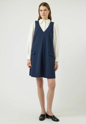 Ženska, oblečena v belo bluzo z dolgimi rokavi pod brezrokavnikom temno modre barve z žepi spredaj, obuta v črne natikače, stoji v nevtralnem položaju.