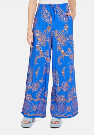 Szerokie niebieskie spodnie z pomarańczowymi wzorami paisley i kwiatowymi, z elastycznym pasem i troczkiem, noszone z jasnoniebieskimi sandałami na platformie.