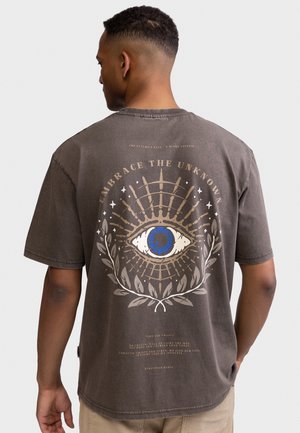 Homme portant un t-shirt marron avec un grand œil bleu et le texte "Embrace the Unknown" dans le dos, debout devant un fond uni.