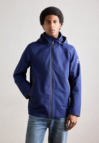 Veste imperméable bleu marine avec capuche, dotée d'une fermeture éclair devant et de deux poches latérales. Fabriquée en tissu lisse et léger avec des accents minimaux.