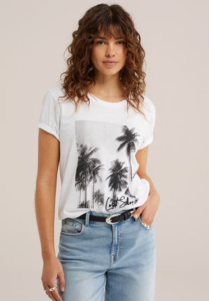 Kvinde med krøllet hår iført en hvid t-shirt med sort palme træ-print og teksten "Lost Shores", kombineret med lyseblå jeans og et sort bælte.
