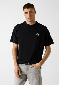 Schwarzes Baumwoll-T-Shirt mit kurzen Ärmeln, rundem Ausschnitt und einem weißen Logo auf der Brust. Kombiniert mit hellgrauen Jeans und einem schwarzen Gürtel.