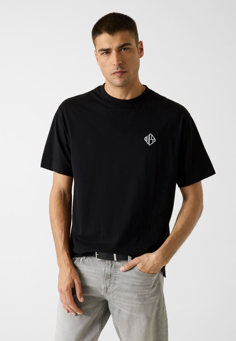 Schwarzes Baumwoll-T-Shirt mit kurzen Ärmeln, rundem Ausschnitt und einem weißen Logo auf der Brust. Kombiniert mit hellgrauen Jeans und einem schwarzen Gürtel.