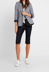Blazer de cuadros grises con un diseño ajustado, combinado con una camiseta gris clara, pantalones cortos de mezclilla oscuros y zapatillas blancas. Bolsa cruzada negra.