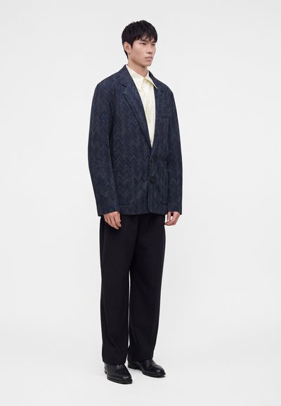 Jeune homme portant un blazer bleu foncé à motif, une chemise claire, un pantalon ample noir et des chaussures noires, debout devant un fond blanc uni.