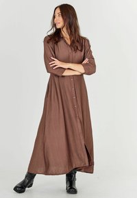 Brown, long-sleeve maxi dress in lightweight fabric with a buttoned front and side slits, paired with black ankle boots.