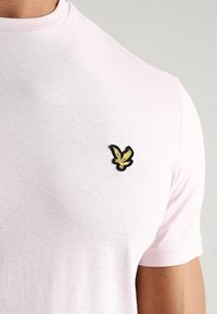 T-shirt rosa chiaro a maniche corte realizzato in un tessuto morbido. Presenta un piccolo logo di un uccello ricamato in nero e oro sul lato sinistro del petto.