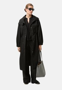 Černá dlouhá bunda s kapucí, s předními kapsami a elastickými manžetami, ve spojení s černým oblečením a světlě šedou taškou přes rameno.