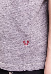 Grått tyg t-shirt med en liten röd broderad logo på vänster sida. Materialet har en strukturerad yta med synliga sömmar.