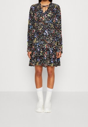 Femme portant une robe noire à manches longues avec des motifs abstraits colorés et des bottines blanches, debout devant un mur clair uni.