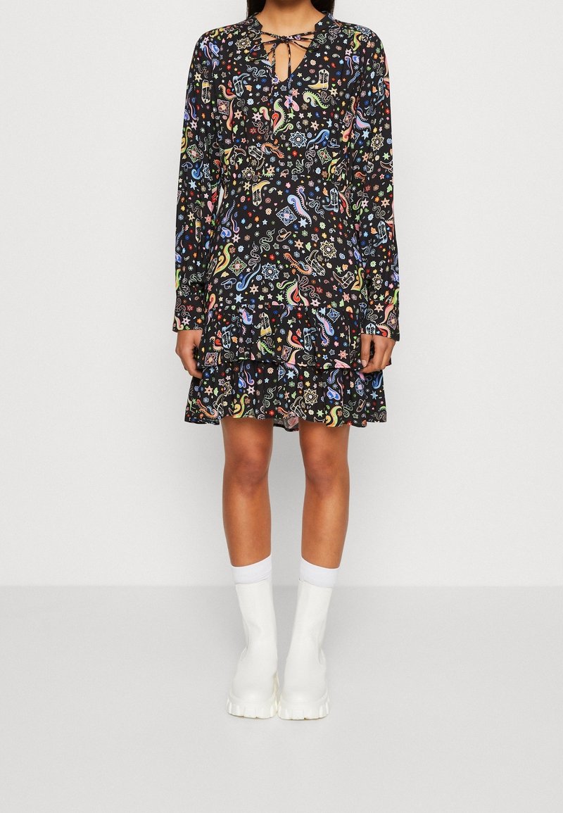 Femme portant une robe noire à manches longues avec des motifs abstraits colorés et des bottines blanches, debout devant un mur clair uni.