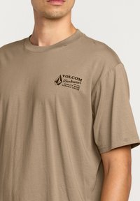 T-shirt à manches courtes de couleur beige avec un logo frontal marron foncé, design col rond et texture en coton doux.