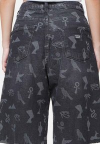 Pantalones cortos de mezclilla negra con un estampado total de símbolos antiguos y animales, con dos bolsillos traseros y una etiqueta en el bolsillo derecho.