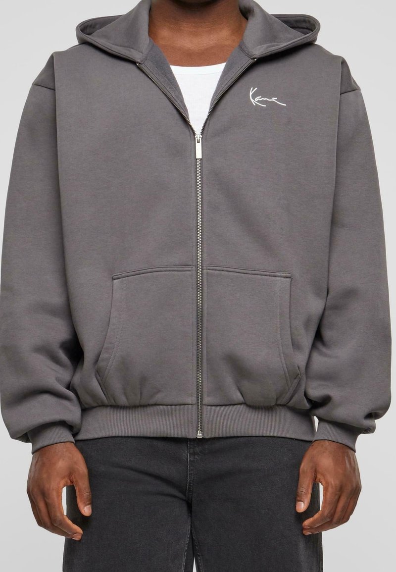 Sudadera con cremallera en gris de tela suave, con un bolsillo tipo canguro, puños acanalados y un pequeño logo blanco en el lado izquierdo del pecho.