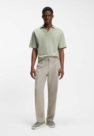 Jonge man met een lichtgroen gebreid shirt met korte mouwen, beige rechtvallende broek en grijze sneakers, staand tegen een witte achtergrond.