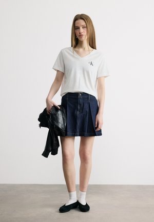 Jauna moteris dėvi baltą V formos kaklo marškinėlį, tamsiai mėlyną džinso sijoną, baltas kojines, juodus batelius ir dešine ranka laiko juodą odinę striukę.